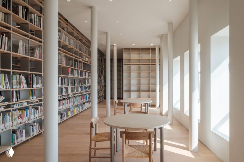 Читальная зона с круглыми столами рядом с высокими окнами и деревянными стеллажами — спокойный уголок для чтения в библиотеке.
