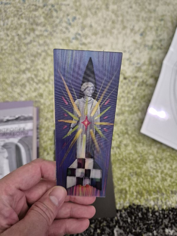 Рука держит яркую двустороннюю закладку с фантастической иллюстрацией, графическими лучами и символикой на тёмном фоне