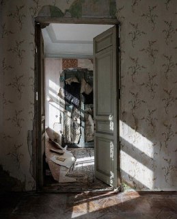 Дверной проём в комнату с облупившимися обоями и накопленными вещами, видны слои старой отделки и следы прежнего быта в коммунальной квартире.