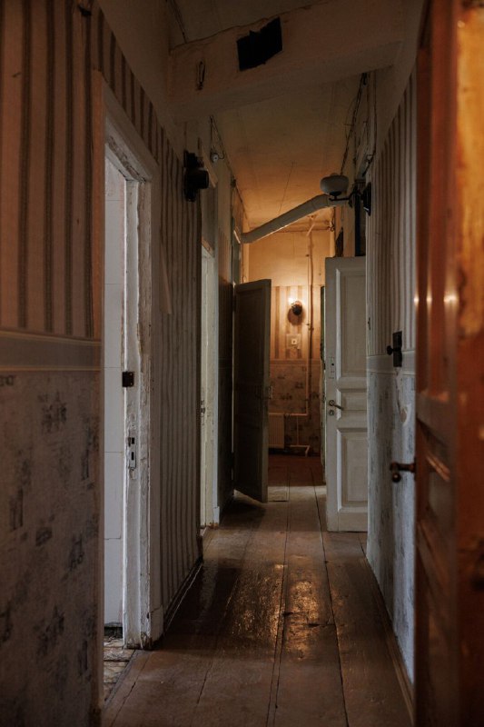 Узкий коридор бывшей коммуналки с потёртым деревянным полом, открытыми дверями и мягким дневным светом, подчёркивающим возраст и атмосферу помещения.