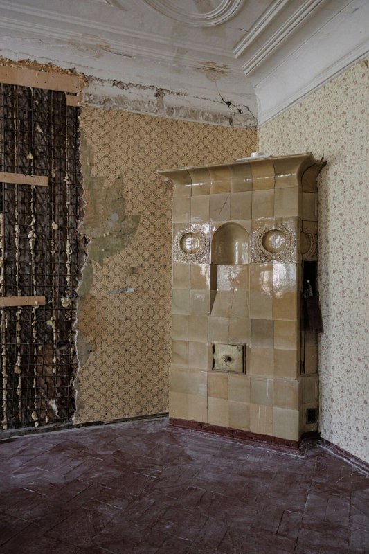 Старинная кафельная печь в комнате с частично отклеившимися обоями и лепниной на потолке — заметный элемент интерьера, сохранённый при расчистке помещения.