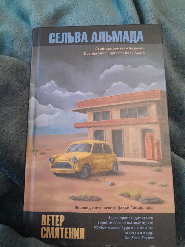 Обложка книги Сельва Альмада с изображением дома и жёлтого автомобиля, книга лежит на текстильном фоне; спокойный кадр для букофила.