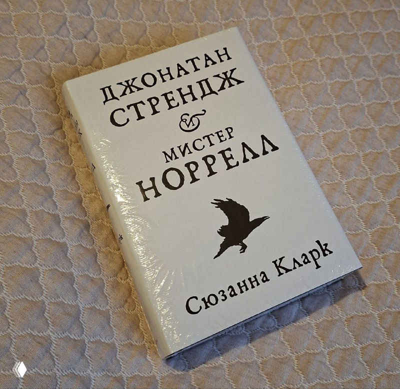 Запечатанная в плёнке книга «Мистер Норрелл» Джонатана Стренджа — белая обложка с силуэтом птицы, снимок крупным планом на тканевом фоне.
