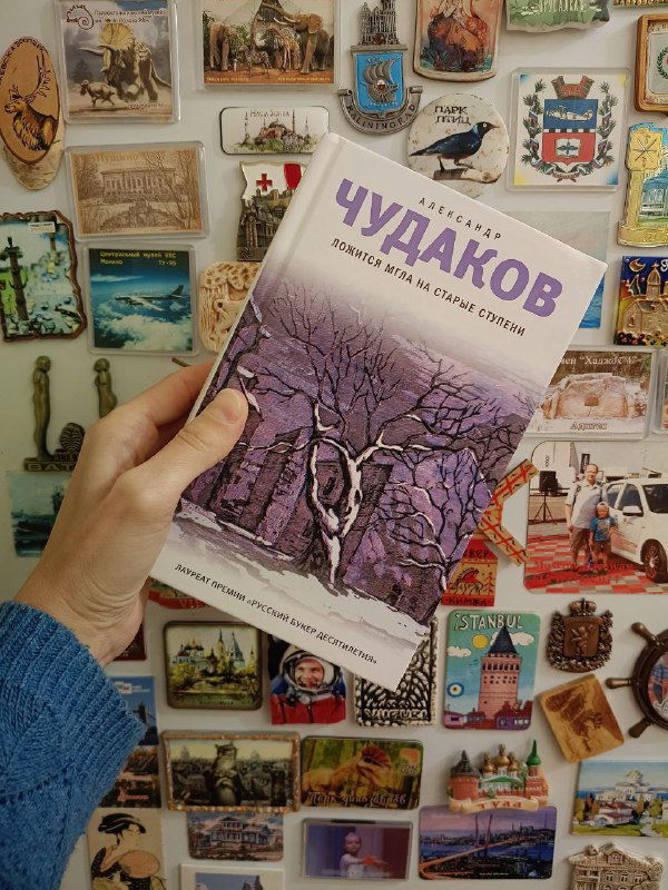 Книга «Чудаков» в руке на фоне коллекции магнитов и сувениров на стене; крупный план обложки с растительным орнаментом.