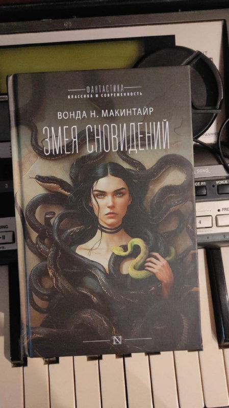 Книга Вонда Н. МакИнтайр «Змея сновидений» лежит на клавишах синтезатора; обложка с портретом и змеёй, творческая студийная атрибутика.