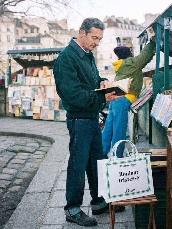 Мужчина читает книгу возле прилавка с книгами, рядом стоит сумка Dior с оформлением обложки книги в зелёной рамке.