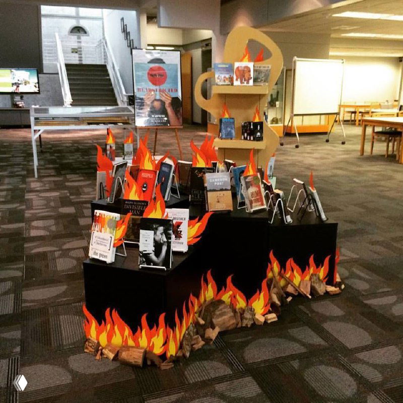 Стеллаж и подиум в библиотеке с книгами, окружёнными декоративными огнями и вырезанными бумажными пламенами — оформление для недели запрещённых книг.