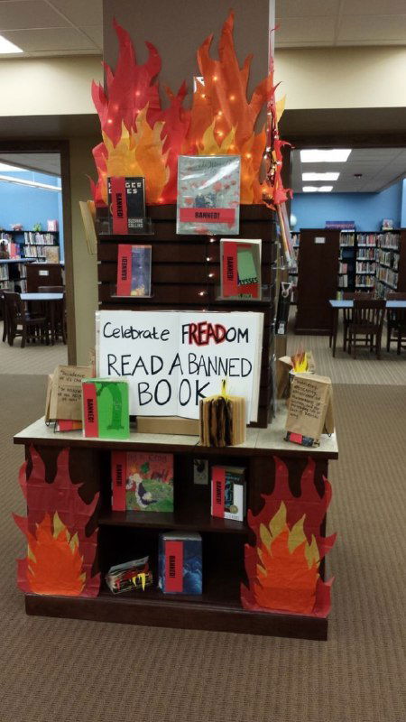 Высокий книжный стенд с надписью и бумажными пламенами; книги выставлены лицом к читателю на фоне читального зала библиотеки.