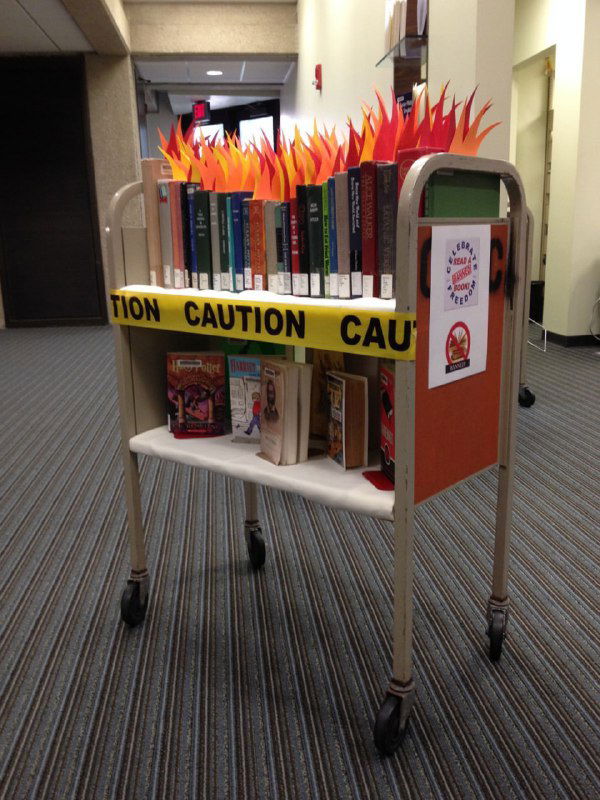 Передвижная тележка с книгами и лентой «CAUTION», украшенная бумажными пламёнами — креативное оформление в библиотеке, привлекающее внимание посетителей.