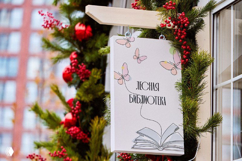 Вывеска «Лесная библиотека» на фоне празднично украшенной ели и гирлянд у входа в небольшое читального павильона в парке.