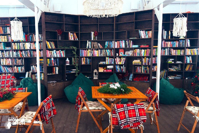 Ещё один ракурс внутреннего пространства: столики, кресла, полки с книгами и тёплое освещение для комфортного чтения.