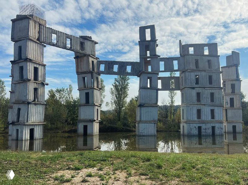 Фотография крупной бетонной инсталляции под открытым небом: высокие железобетонные модули и отражение в воде, архитектурная руина-монумент.