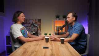 Павликов и ведущий разговаривают в подкаст-студии: два человека за деревянным столом у микрофонов, на столе чашки и смартфон, мягкая подсветка на фоне.