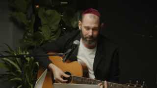 Исполнитель в сумраке с акустической гитарой у микрофона, сценический портрет, иллюстрирующий тему пародии на образ «шаман» в тексте.