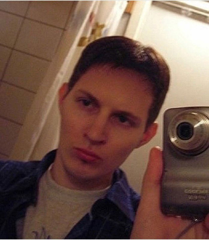 Зеркальное селфи с камерой в кадре: молодой человек держит цифровой фотоаппарат, видна плитка ванной и бытовой фон.