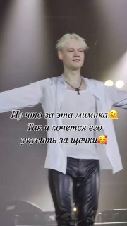 Артист на сцене в белой рубашке и кожаных штанах, руки в стороны, сценические прожекторы и текстовый субтитр на кадре, кадр из концертного видео.