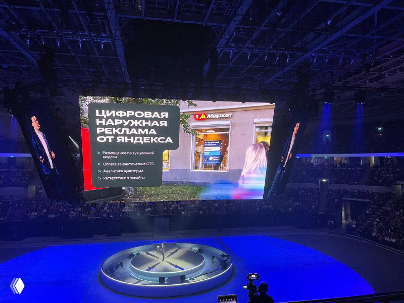 Презентация цифровой наружной рекламы от Яндекса на большом экране: пример баннера, сцена с подсветкой и публика внизу кадра.