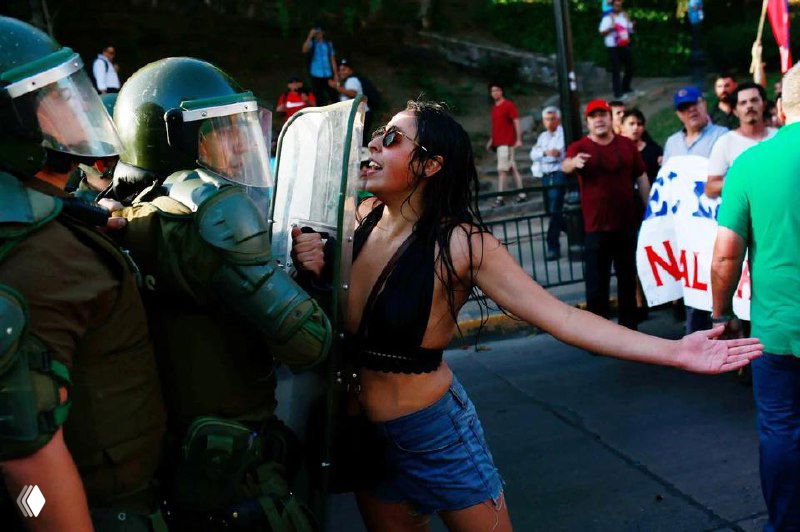 Молодая женщина в провокационной позе перед линией полицейских в касках и щитах — напряжённый момент конфронтации на улице.