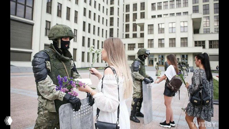 Группа молодых женщин общается с солдатами в форме у административного здания; цветы и сумки усиливают контраст между ними.