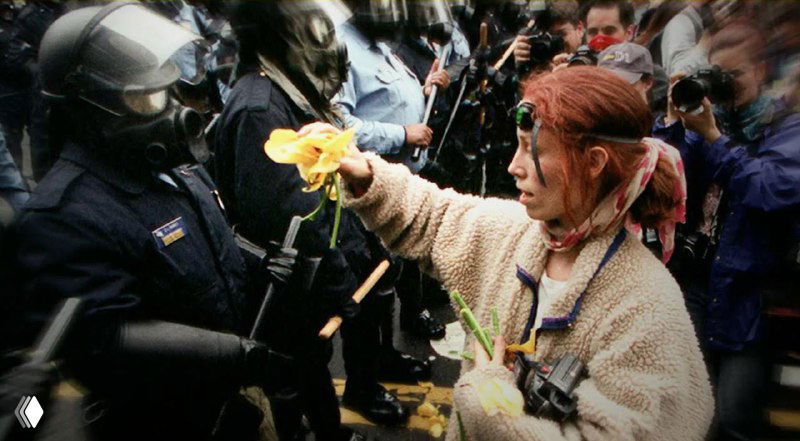 Пожилая женщина протягивает цветок скоплению полицейских в касках; кадр передаёт эмоциональную попытку пацифистского контакта.