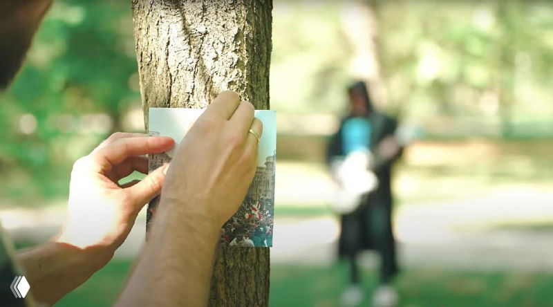 Рука прикалывает листовку к коре дерева в парке, на заднем плане размыто виден человек с гитарой — кадр с призывом расклеивать листовки.