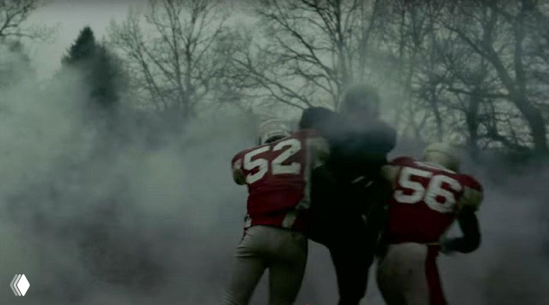 Крупный план двух игроков в красных номерах и темном сопернике в дымке — образ насилия и конфронтации, использованный в клипе для метафоры протеста.