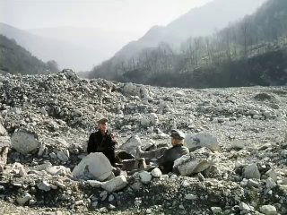 Кадр из фильма «Свой среди чужих»: двое мужчин сидят среди крупных камней в широком каменистом русле реки, на фоне туманных гор и обрывистых склонов.