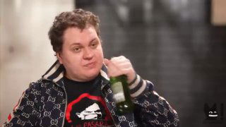 Крупный план кадра из видео: мужчина в куртке с принтом держит бутылку в руке в неформальной обстановке, напряжённая миниатюрная сцена беседы.