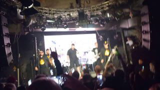 Размытое фото концертной сцены с Сергеем Михалком и музыкантами, публика в тёмном зале снимает выступление на телефоны и держит руки вверх.