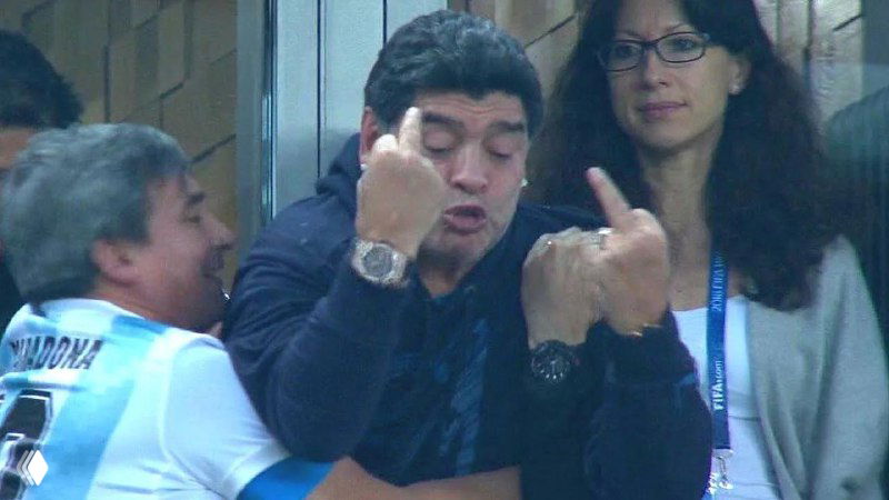 Диего Марадона на трибуне показывает неприличные жесты среди болельщиков, снимок с футбольного матча с выраженной эмоцией и реакцией людей рядом.