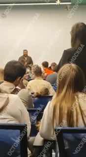 Зал с аудиторией студентов на встрече: спины слушающих, сцена впереди и лектор у белой стены; виден водяной знак по краю кадра.