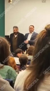 Два человека у стены перед аудиторией на встрече: режиссёр и организатор стоят у белой стены; видны сидящие студенты и водяной знак.