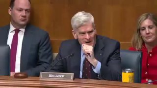 Заседание в зале Сената: мужчина в костюме у микрофона говорит за столом на слушаниях, вокруг другие участники и таблички на столах, ситуация официальная