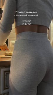 Кадр сзади: женщина в обтягивающих светло‑серых леггинсах готовит на кухне; поверх фото видна надпись с рецептом и КБЖУ.