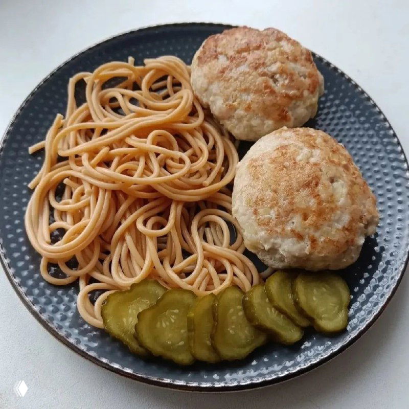 Тёмная тарелка со спагетти, двумя куриными котлетками и ломтиками солёных огурцов — простой ПП-обед, снят сверху на кухонном столе.