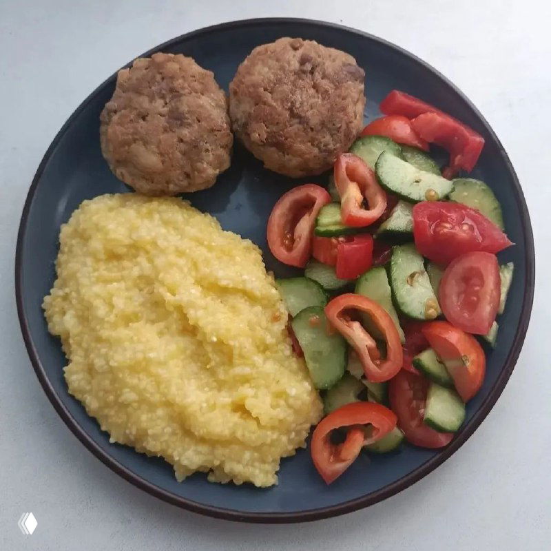 Тарелка с кукурузной кашей, двумя котлетами и овощным салатом из помидоров и огурцов — классический ПП-обед, вид сверху на тёмной посуде.