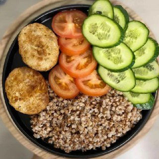 Тарелка с варёной гречкой, ломтиками огурца и помидора и двумя котлетами или котлетками — простой и сбалансированный ПП-обед, вид сверху.