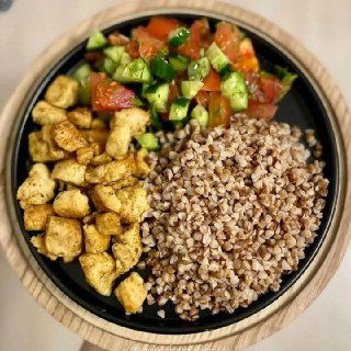 Тарелка с гречкой и кусочками обжаренной грудки, рядом овощной салат — порция ПП-обеда на деревянной подставке, вид сверху.