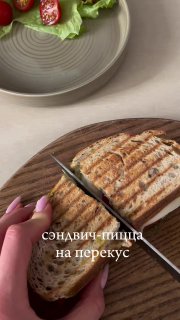 Разрезанный тёплый сэндвич на деревянной разделочной доске; на заднем плане тарелка с салатом и помидором — иллюстрация сытного перекуса из поста.