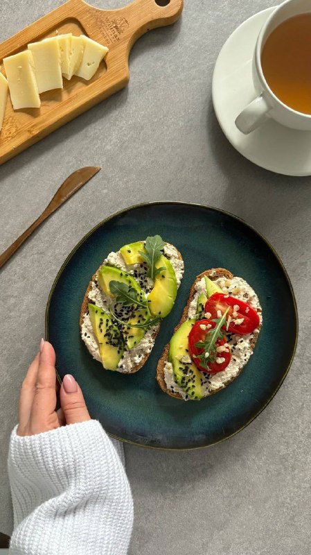 Два тоста на тёмной тарелке с ломтиками авокадо, творогом и посыпкой семян, рядом чашка напитка — эстетичная ПП-подача.