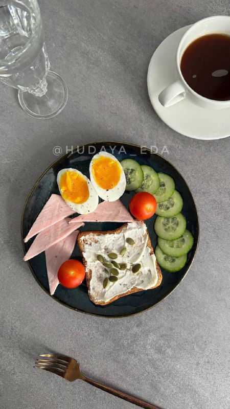 Тарелка с тостом, творожным сыром, ломтиками яйца, карбонатом и овощами; рядом кружка чая и огурцы — сбалансированный приём пищи.