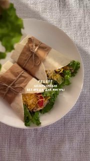 Сверху: цезарь-ролл, завёрнутый в пергамент и перевязанный ниткой, на белой тарелке с листами салата, дольками помидора и огурца.