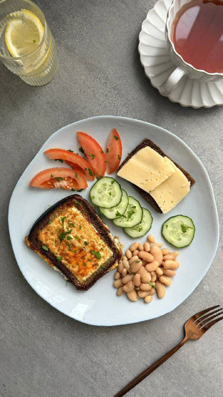 Тарелка с тостом-яичницей в хлебе, порцией фасоли, ломтиками томата и кусочком лёгкого сыра; рядом чашка тёплого напитка и вилка на столе.