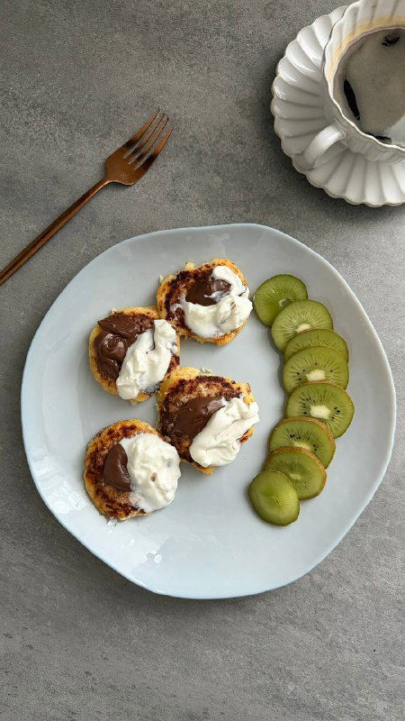 Порция сырников из обезжиренного творога, политы греческим йогуртом и шоколадным соусом, рядом нарезанный киви и минималистичная сервировка на белой тарелке.