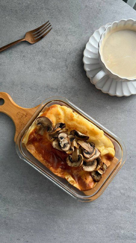 Запечённый омлет с грибами в прямоугольной стеклянной форме, сверху жареные грибы, рядом чашка тёплого напитка — простая домашняя сервировка.