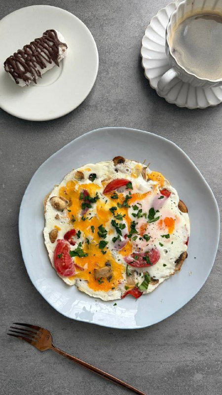 Тарелка с яичницей, обжаренными грибами и помидорами, украшенная зеленью, рядом ложка и минимум аксессуаров — домашний ПП-завтрак на тарелке.