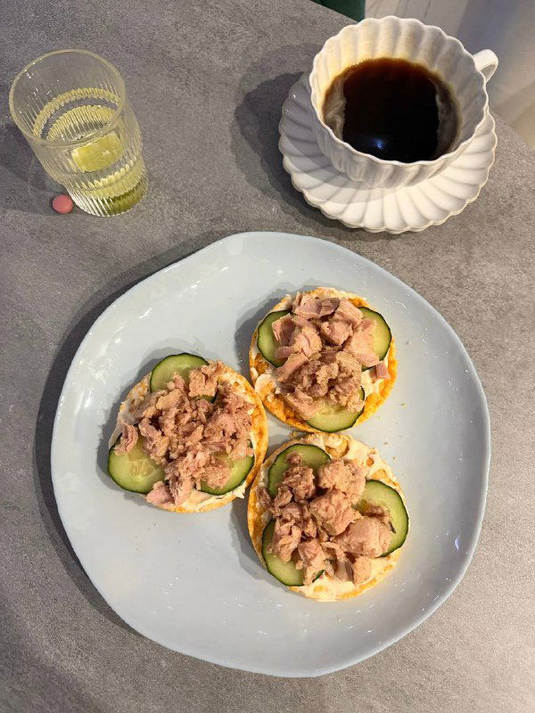 Три хлебца с ломтиками огурца и консервированным тунцом на тарелке, рядом стакан с напитком и чашка кофе — вариант с тунцом и огурцом.