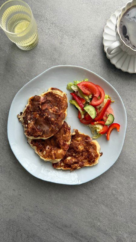 Тарелка с оладьями из творога, рядом свежий овощной салат и чашка чая на сером столе — сытный низкокалорийный вариант.