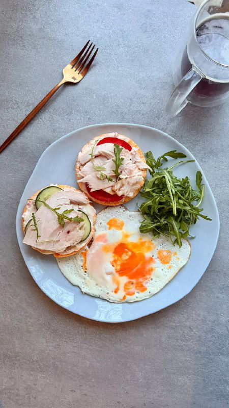 Тарелка с яйцом, ломтиком хлебца с сыром и кусочками куриной грудки, рядом руккола и стакан воды — сбалансированный белковый завтрак.