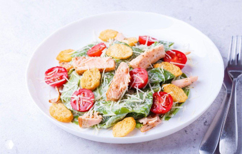 Салат с кусочками рыбы на гриле, зеленью и помидорами черри, сверху хрустящие гренки, подача в неглубокой белой тарелке.